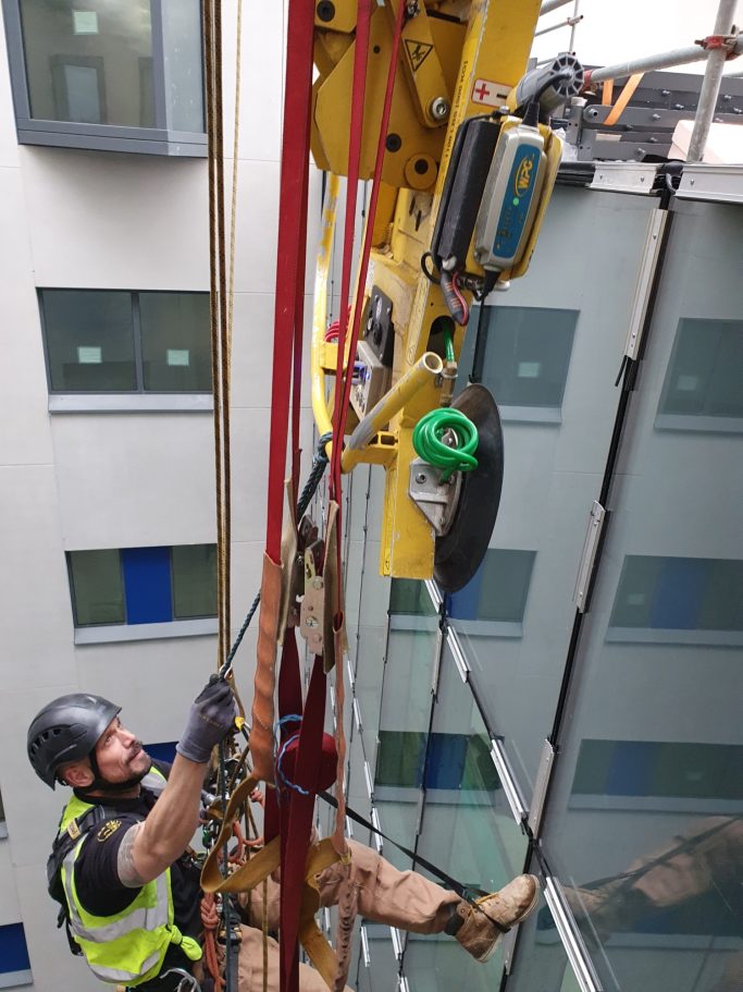 Worker in safety gear using a rope access system on a building's glass façade.
