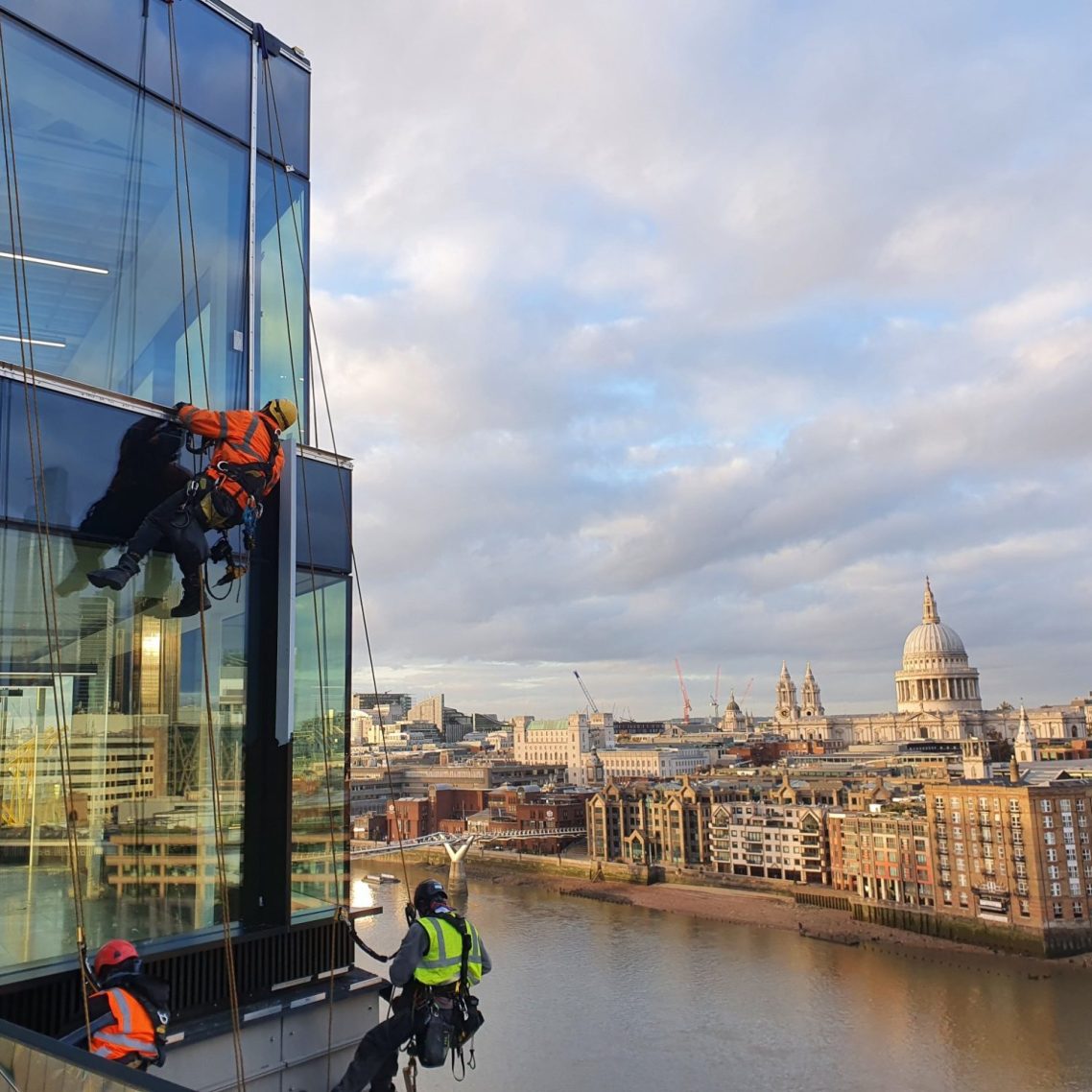 Workers abseiling down a glass building with St Paul's Cathedral and the river Thames in the background.