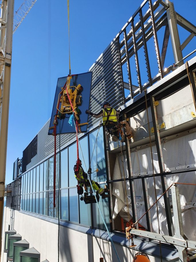 Workers using ropes to install a large glass panel on a building's façade.